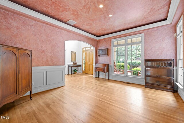 empty room featuring arched walkways, crown molding, light wood finished floors, a wainscoted wall, and a tray ceiling