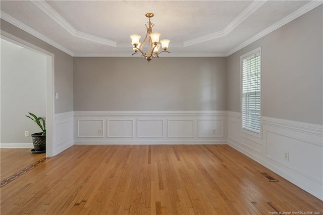 empty room with a chandelier, light wood finished floors, wainscoting, crown molding, and a raised ceiling