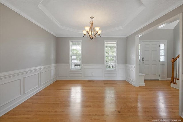 unfurnished dining area with a chandelier, light wood-type flooring, a wainscoted wall, a tray ceiling, and ornamental molding