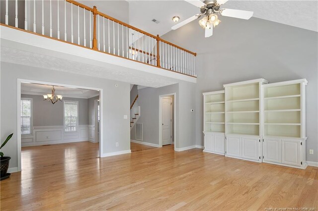 unfurnished living room with stairs, a chandelier, ceiling fan, light wood-style flooring, and a towering ceiling