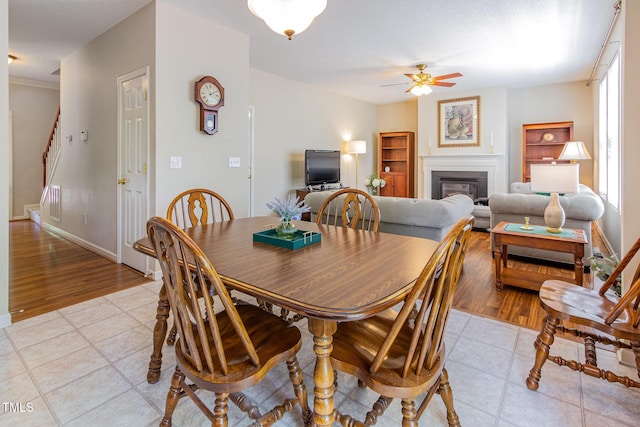 dining space with ceiling fan, light tile patterned floors, a glass covered fireplace, stairs, and baseboards