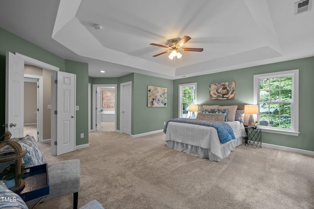 bedroom with a tray ceiling, baseboards, light colored carpet, and ensuite bath