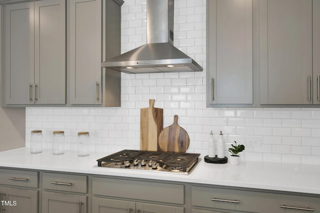 kitchen featuring gray cabinetry, wall chimney range hood, stainless steel gas stovetop, light countertops, and tasteful backsplash