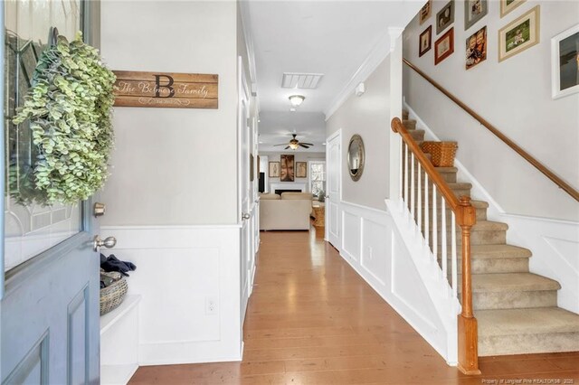foyer entrance with wood finished floors, crown molding, stairway, a ceiling fan, and wainscoting