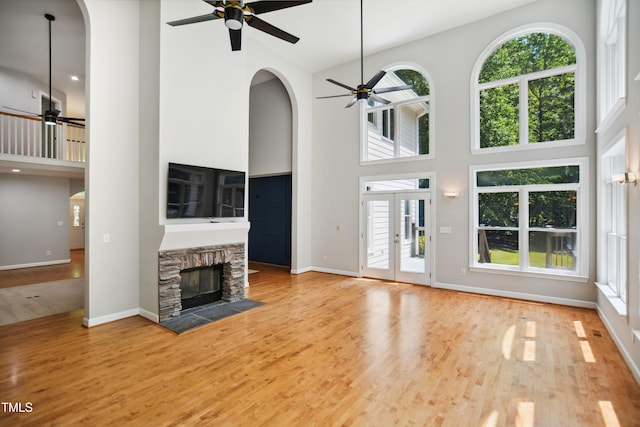 unfurnished living room featuring a ceiling fan, a high ceiling, a stone fireplace, wood finished floors, and baseboards