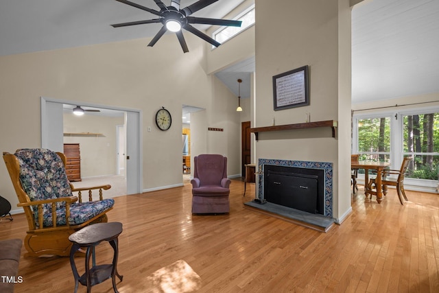 living area featuring a tiled fireplace, a ceiling fan, light wood-type flooring, high vaulted ceiling, and baseboards