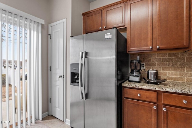 kitchen with stainless steel refrigerator with ice dispenser, brown cabinets, backsplash, and light stone countertops