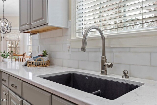 kitchen view of a sink, tasteful backsplash, gray cabinetry, light stone countertops, and a chandelier