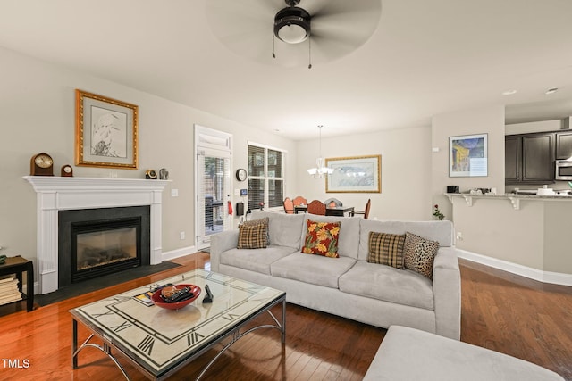 living room featuring ceiling fan, wood finished floors, a fireplace with flush hearth, a chandelier, and baseboards