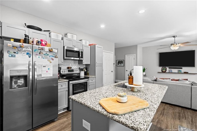 kitchen with appliances with stainless steel finishes, wood finished floors, a kitchen island with sink, a ceiling fan, and light stone counters