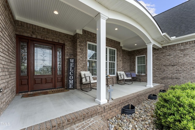 doorway to property featuring a porch, brick siding, and roof with shingles