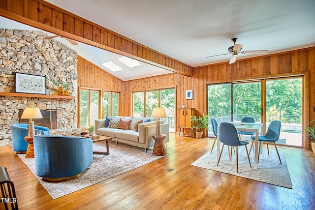 living room featuring wooden walls, a ceiling fan, a skylight, wood finished floors, and a stone fireplace