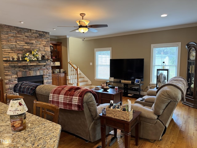 living area with stairs, ceiling fan, wood finished floors, a stone fireplace, and crown molding