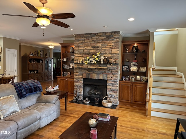 living room with a ceiling fan, crown molding, light wood-type flooring, stairs, and a stone fireplace