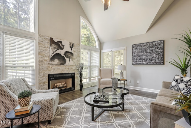 living area with high vaulted ceiling, wood finished floors, ceiling fan, a stone fireplace, and baseboards