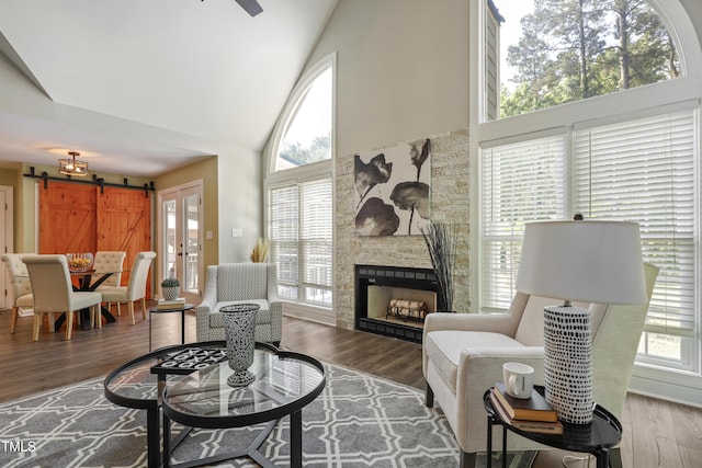 living area with high vaulted ceiling, healthy amount of natural light, wood finished floors, a barn door, and a stone fireplace