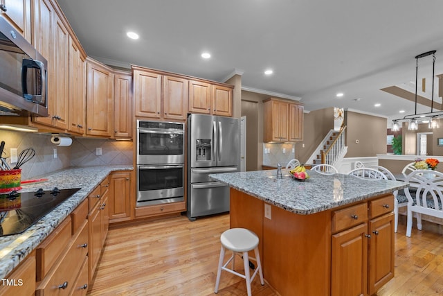 kitchen with appliances with stainless steel finishes, ornamental molding, light wood finished floors, a center island with sink, and light stone countertops