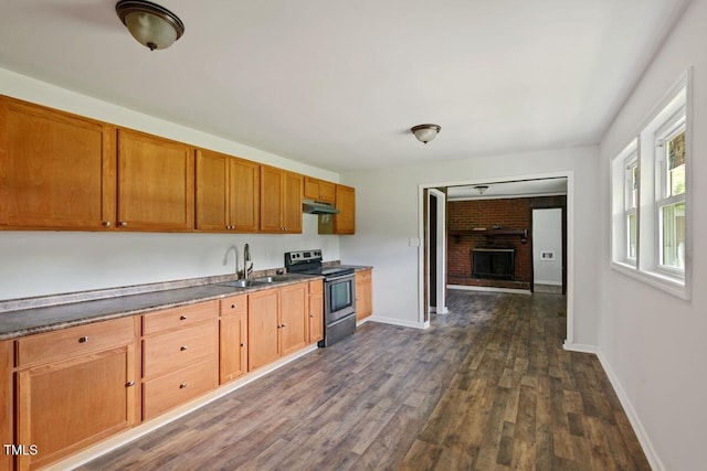 kitchen with stainless steel electric range oven, a sink, under cabinet range hood, dark wood-type flooring, and a brick fireplace