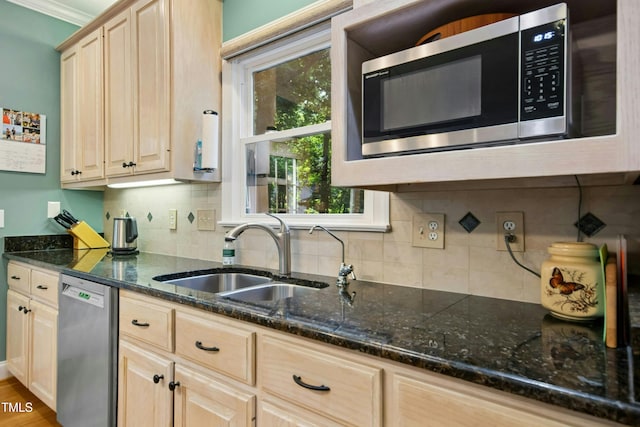 kitchen with stainless steel appliances, a sink, light brown cabinets, backsplash, and dark stone countertops