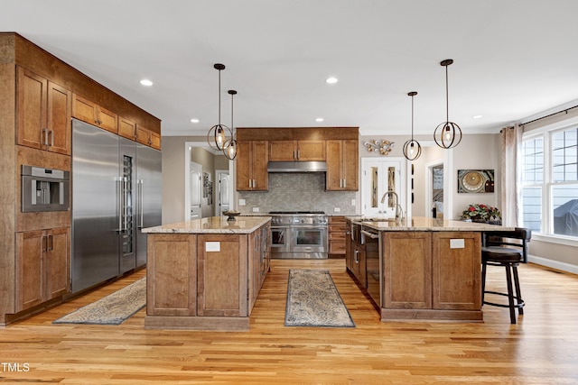 kitchen with high quality appliances, an island with sink, under cabinet range hood, brown cabinetry, and light wood finished floors