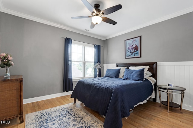 bedroom featuring light wood-style flooring, ornamental molding, a wainscoted wall, a ceiling fan, and baseboards