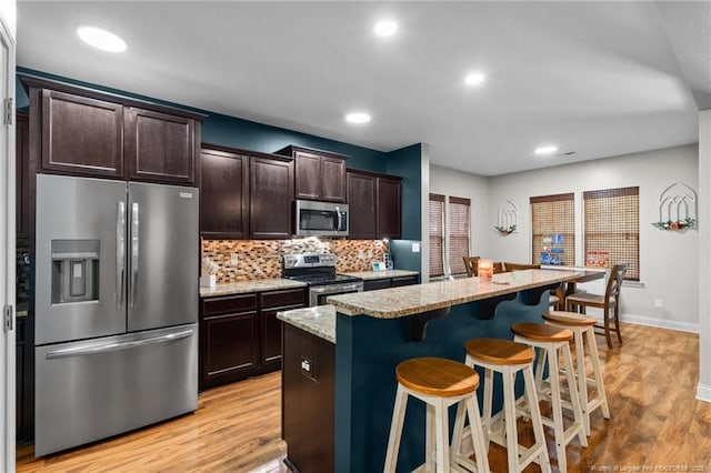 kitchen with appliances with stainless steel finishes, a kitchen breakfast bar, light wood finished floors, dark brown cabinets, and light stone counters