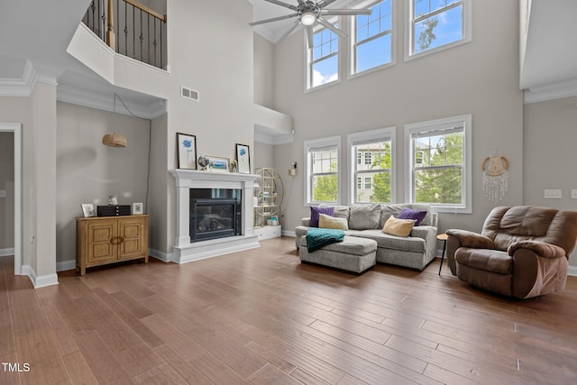 living room featuring ceiling fan, a towering ceiling, crown molding, a glass covered fireplace, and wood finished floors