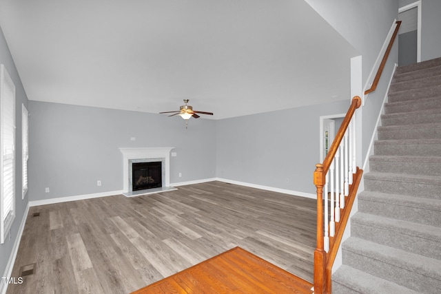 unfurnished living room with ceiling fan, wood finished floors, stairs, baseboards, and a fireplace with raised hearth
