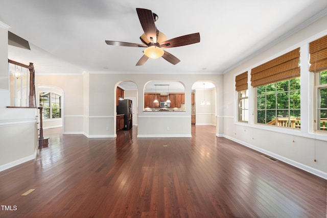unfurnished living room featuring arched walkways, baseboards, dark wood-style floors, ceiling fan, and crown molding
