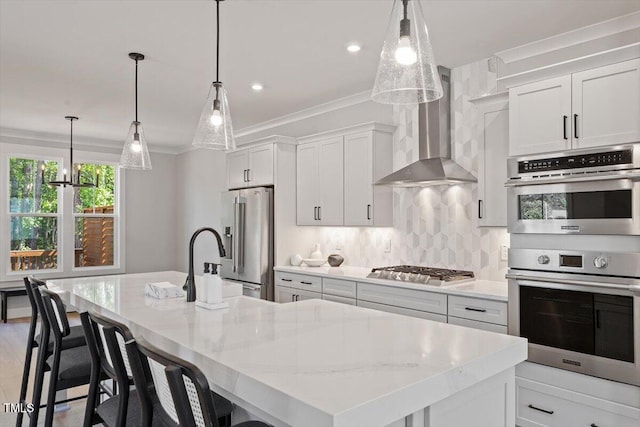 kitchen with stainless steel appliances, wall chimney exhaust hood, ornamental molding, a center island with sink, and backsplash