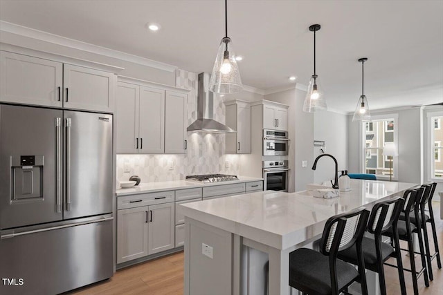 kitchen featuring appliances with stainless steel finishes, wall chimney range hood, a sink, a kitchen island with sink, and ornamental molding