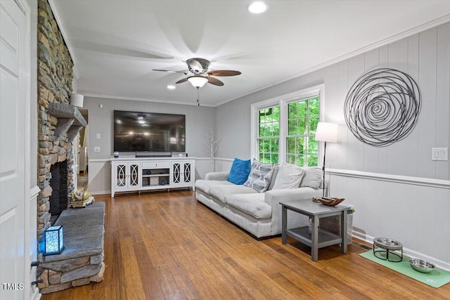 living room featuring ceiling fan, wood finished floors, crown molding, and a stone fireplace