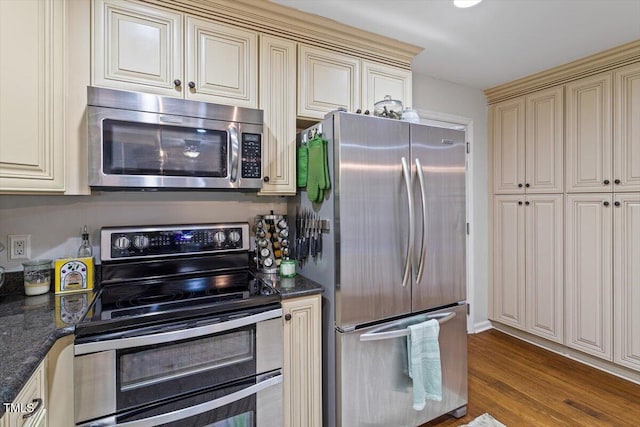 kitchen with appliances with stainless steel finishes, cream cabinetry, wood finished floors, and dark stone countertops
