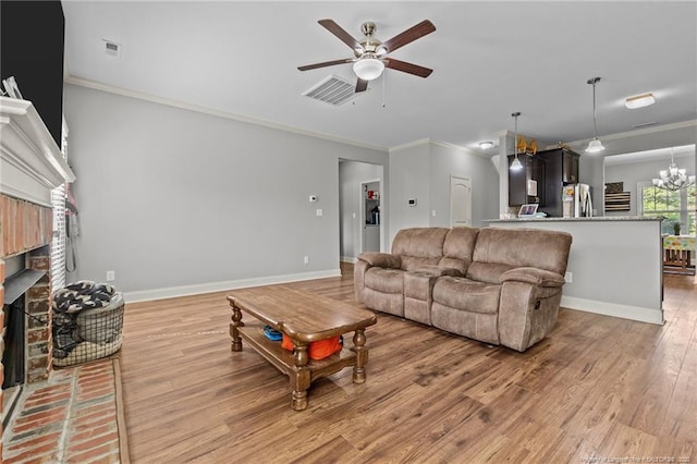 living room featuring a ceiling fan, light wood-type flooring, crown molding, a brick fireplace, and a chandelier