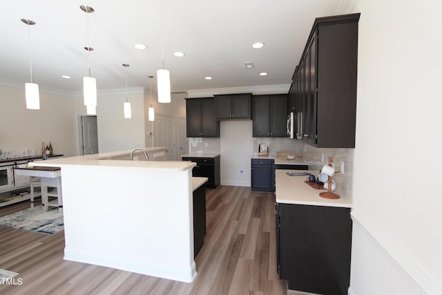 kitchen featuring light wood-style floors, backsplash, a kitchen island with sink, light countertops, and ornamental molding