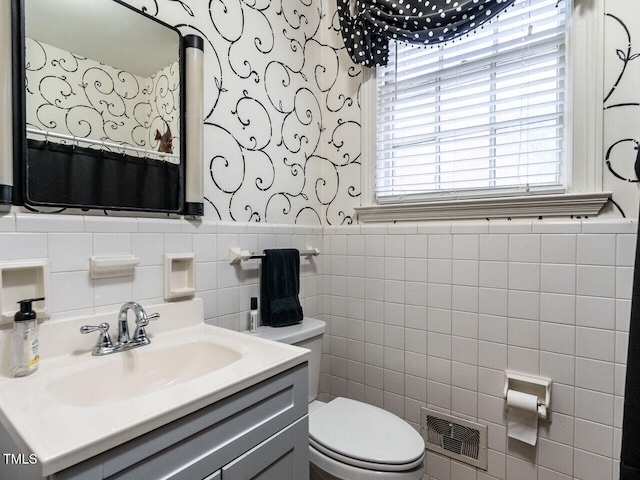 bathroom featuring wallpapered walls, toilet, tile walls, wainscoting, and vanity