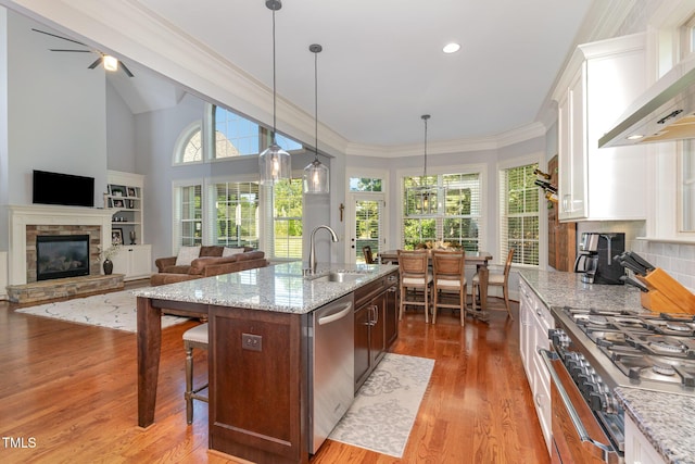 kitchen featuring a sink, a stone fireplace, appliances with stainless steel finishes, wall chimney exhaust hood, and white cabinets