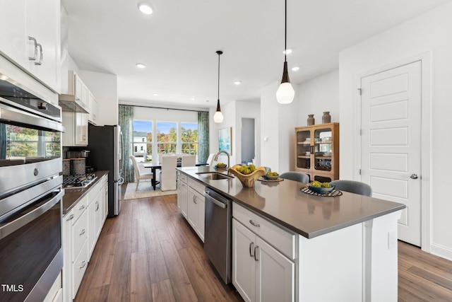 kitchen with stainless steel appliances, a sink, dark countertops, an island with sink, and dark wood-style flooring