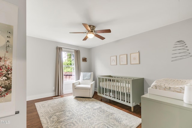 bedroom featuring a nursery area, dark wood finished floors, baseboards, and a ceiling fan