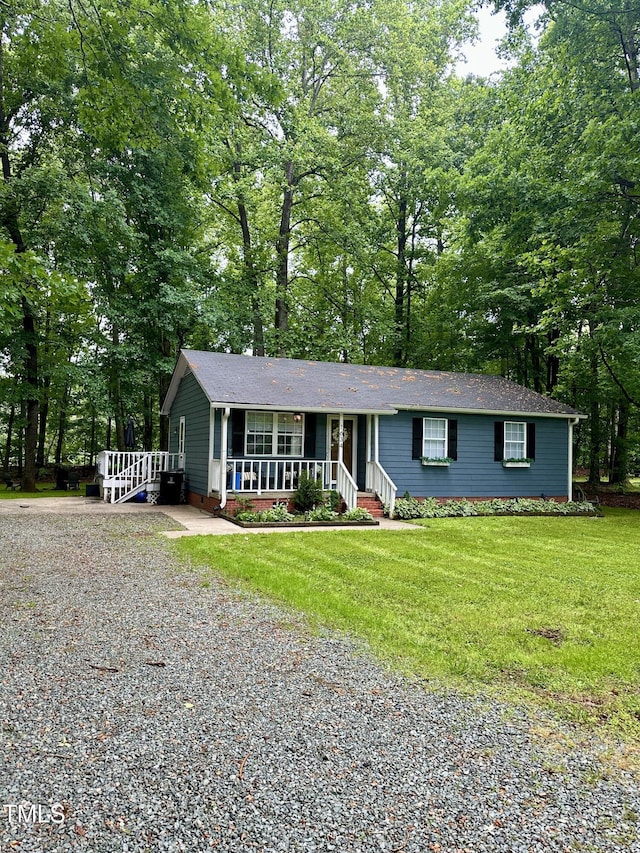 ranch-style house featuring gravel driveway, a porch, a front yard, a shingled roof, and view of wooded area