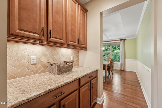 kitchen with wood finished floors, tasteful backsplash, brown cabinets, light stone counters, and a wainscoted wall