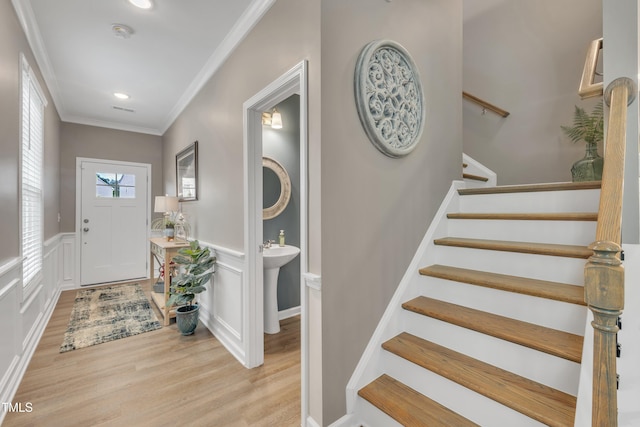 entryway featuring crown molding, stairway, light wood finished floors, a decorative wall, and a wainscoted wall