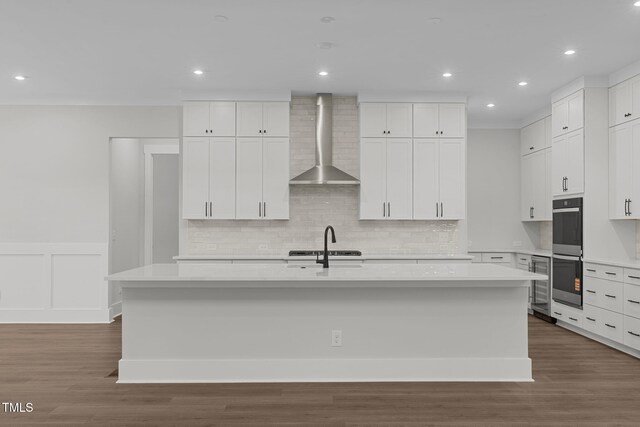 248 Ashford Dr, Pittsboro, NC 27312 - kitchen featuring wall chimney exhaust hood, multiple ovens, a kitchen island with sink, light countertops, and a wainscoted wall
