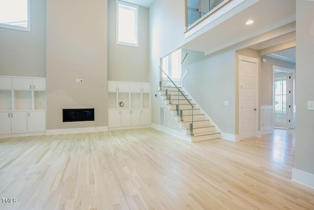 unfurnished living room featuring light wood-style floors, stairs, a glass covered fireplace, a towering ceiling, and baseboards
