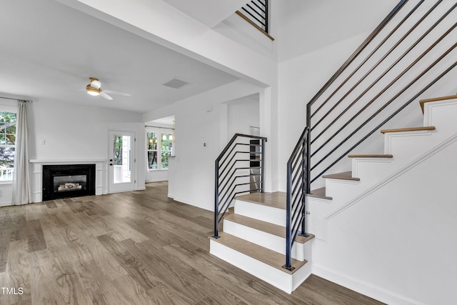 staircase with wood finished floors, ceiling fan, baseboards, and a glass covered fireplace