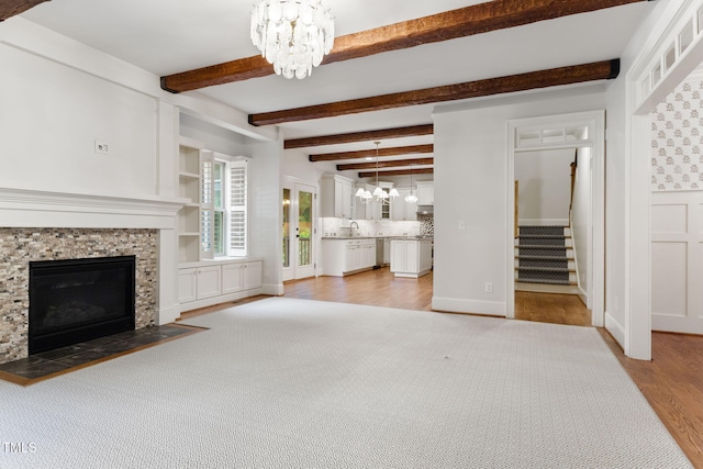 unfurnished living room with a chandelier, light wood-style flooring, a stone fireplace, stairs, and beamed ceiling