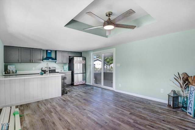 kitchen with wall chimney exhaust hood, stainless steel appliances, a ceiling fan, dark wood finished floors, and light countertops