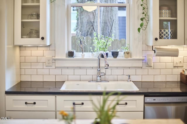 kitchen with stainless steel dishwasher, dark countertops, a sink, white cabinetry, and glass insert cabinets
