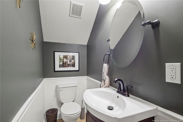 bathroom featuring wainscoting, a decorative wall, toilet, vanity, and lofted ceiling