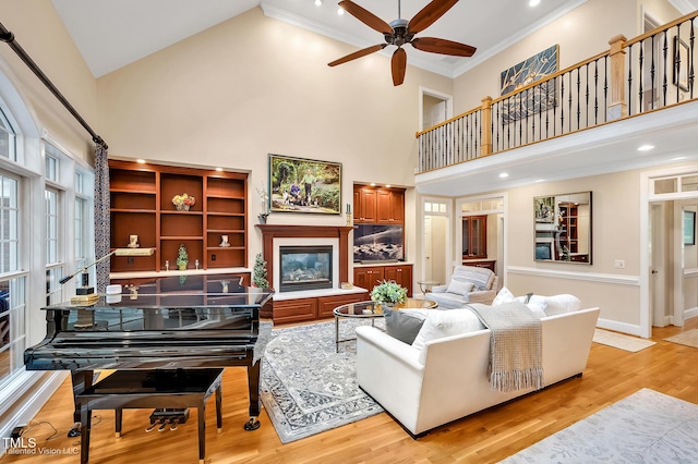 living area with high vaulted ceiling, a glass covered fireplace, ceiling fan, light wood-style flooring, and ornamental molding